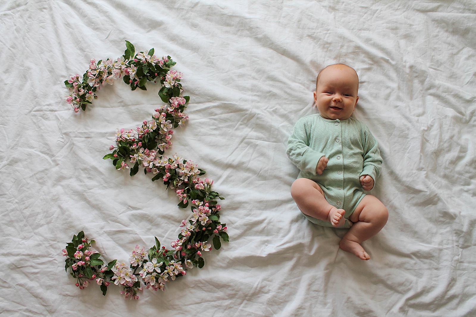 A baby lays beside a large letter three made out of flowers for a newborn month by month picture.