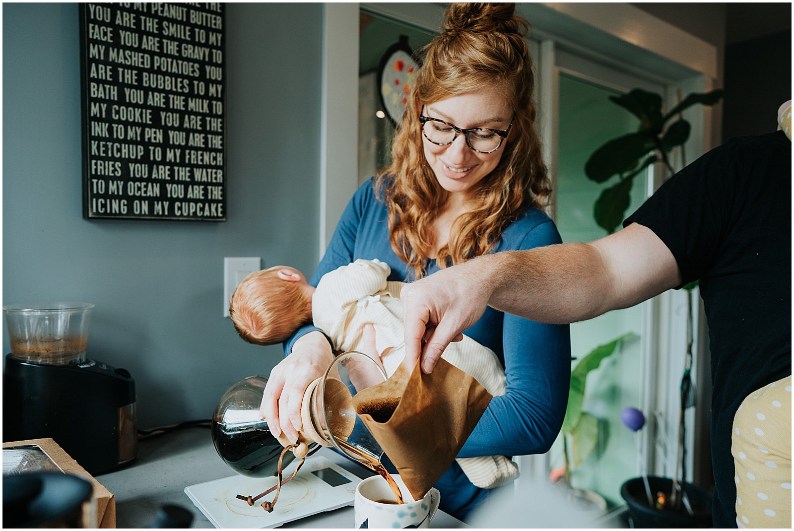 A mother holds a newborn baby while she pours a cup of coffee.