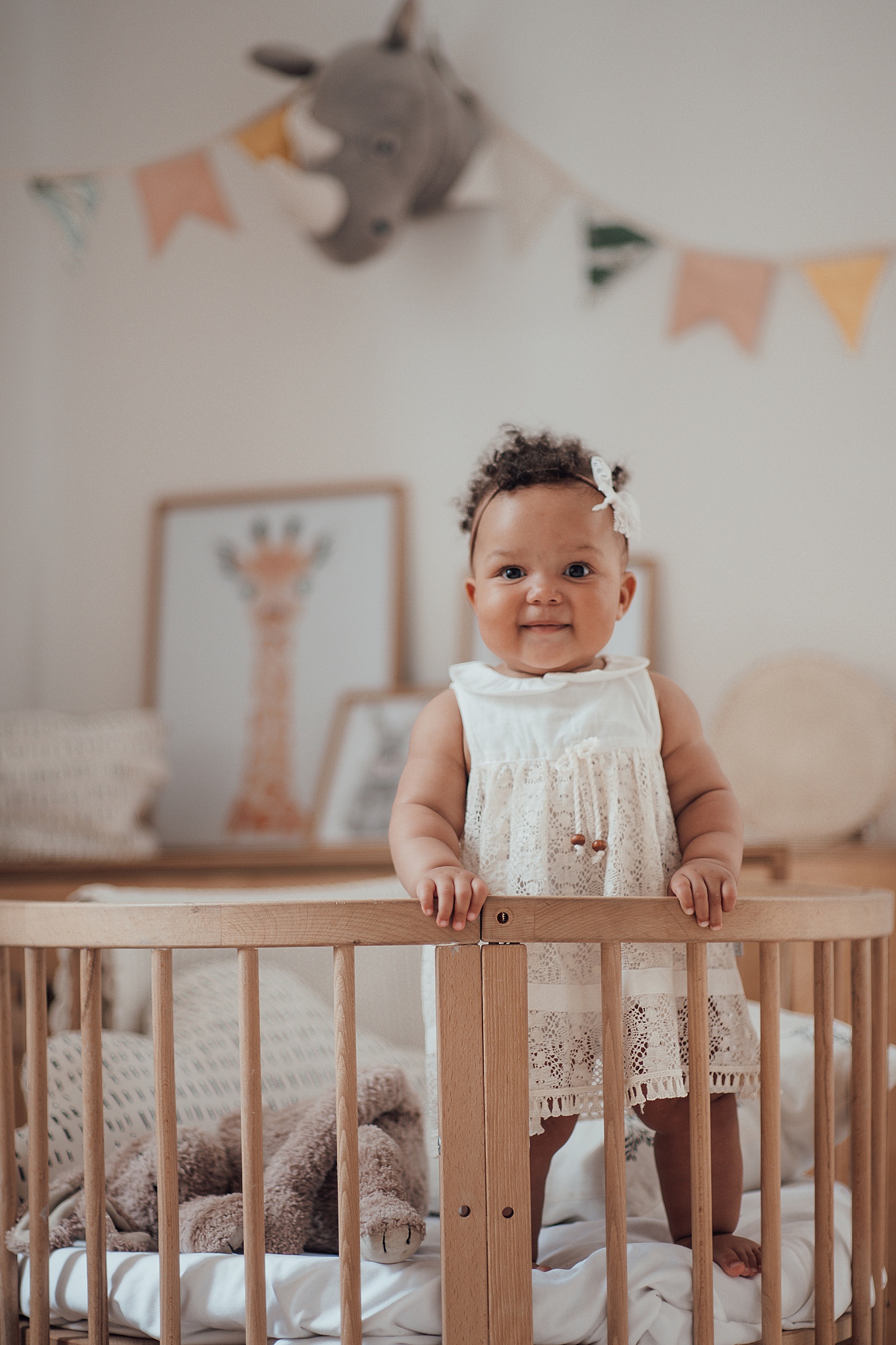 A baby girl stands holding onto the rail of her crib for a monthly milestone photo for her baby book.