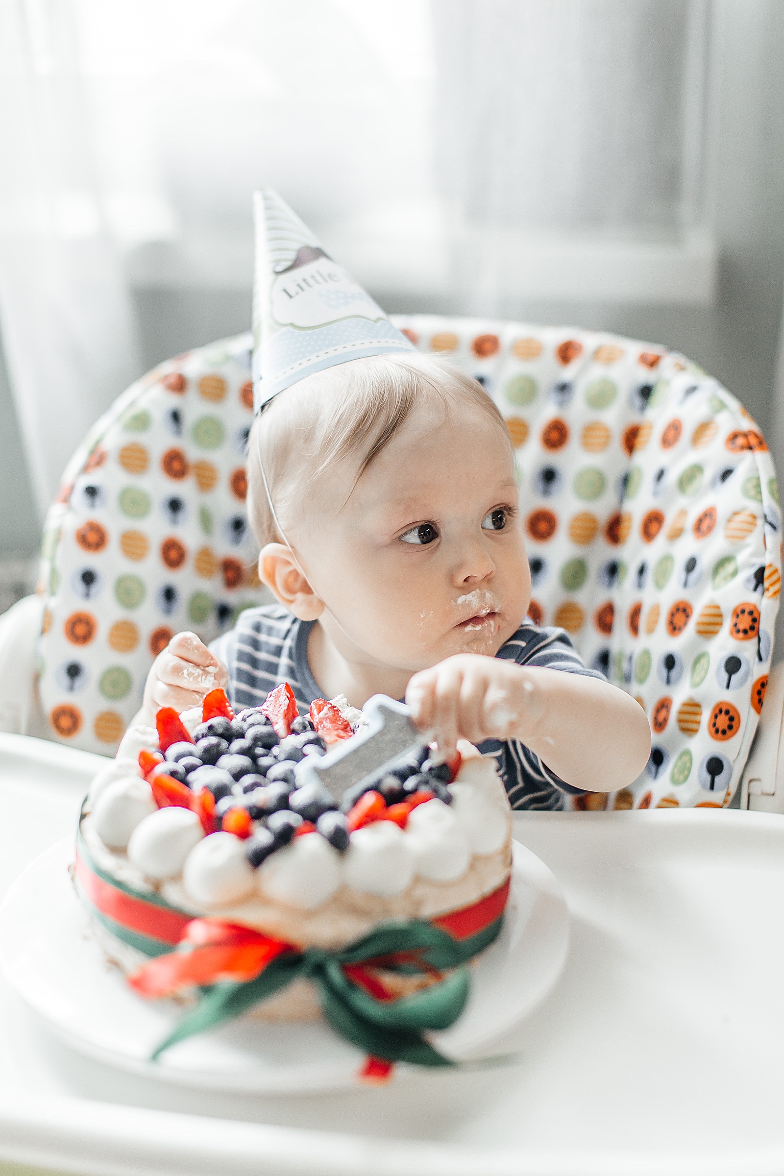 A baby sits in a high chair eating a 1st birthday cake decorated with berries.